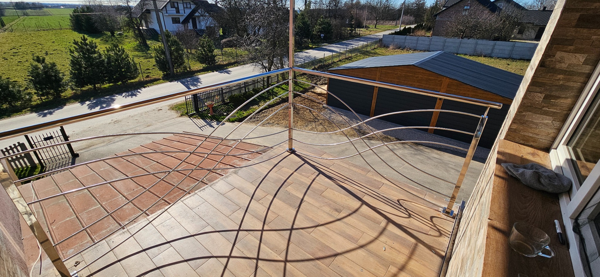 Błyszcząca, metalowa balustrada balkonowa z dekoracyjnymi, falistymi elementami, widziana z góry, z widokiem na domy i ulicę w tle.