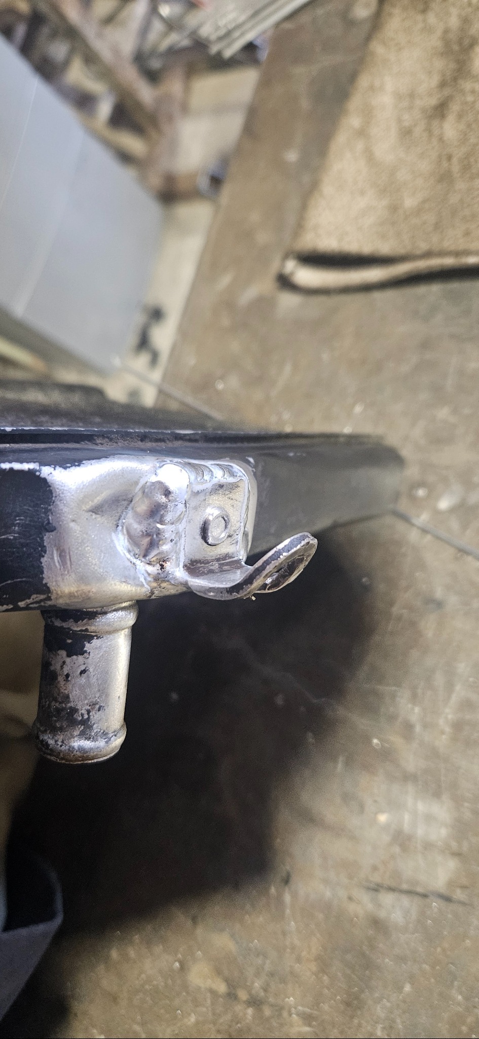 Szczegół spawu na metalowym elemencie z uchwytem, widoczne ślady obróbki i niedoskonałości spoiny. Powierzchnia metalu jest częściowo pomalowana na czarno.