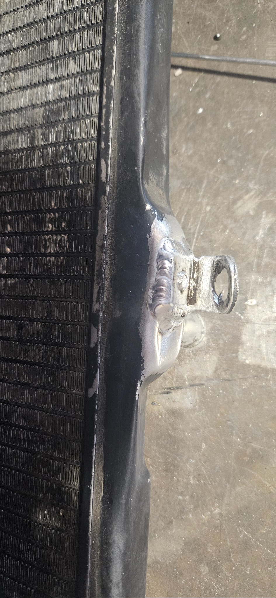 Detal spawu na czarnym elemencie metalowym, widoczne ślady spawania i chropowata powierzchnia. Element przypomina fragment chłodnicy lub innego urządzenia.