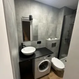 Nowoczesna łazienka w szarościach z drewnopodobną podłogą, umywalką nablatową na czarnym blacie nad pralką, kabina prysznicowa z czarnymi ramami i wisząca toaleta.