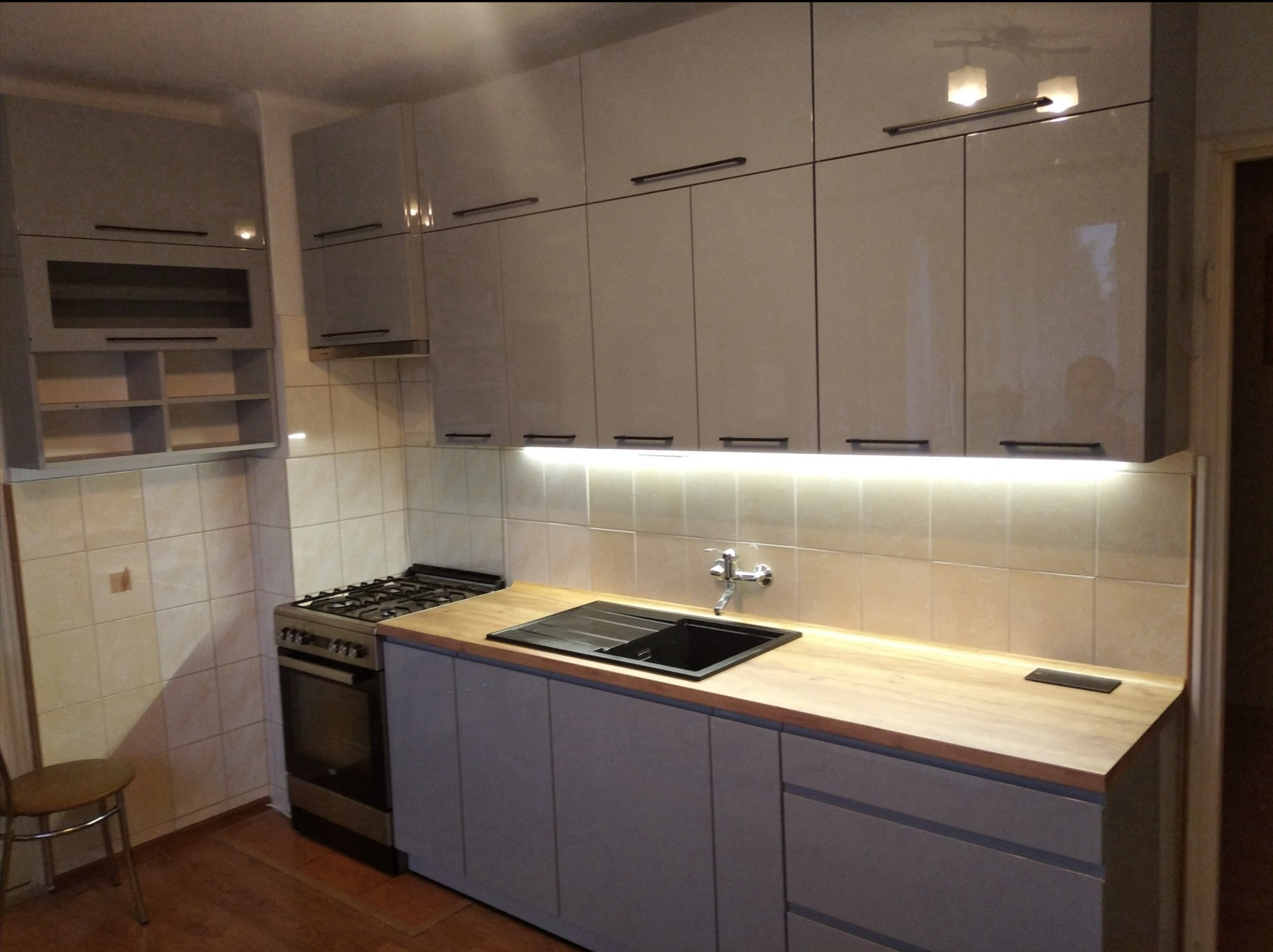 Szara zabudowa kuchenna z drewnianym blatem, czarnym zlewem i płytą gazową, oświetlona listwą LED pod szafkami wiszącymi, widok na fragment wnętrza z krzesłem.