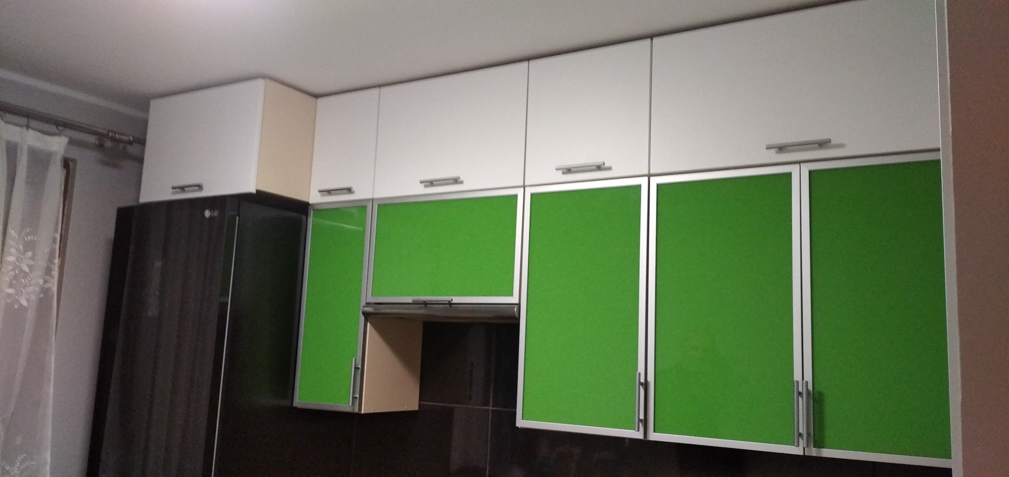 Białe szafki kuchenne wiszące z zielonymi frontami, srebrne uchwyty, obok czarna lodówka LG, ciemne płytki na ścianie.