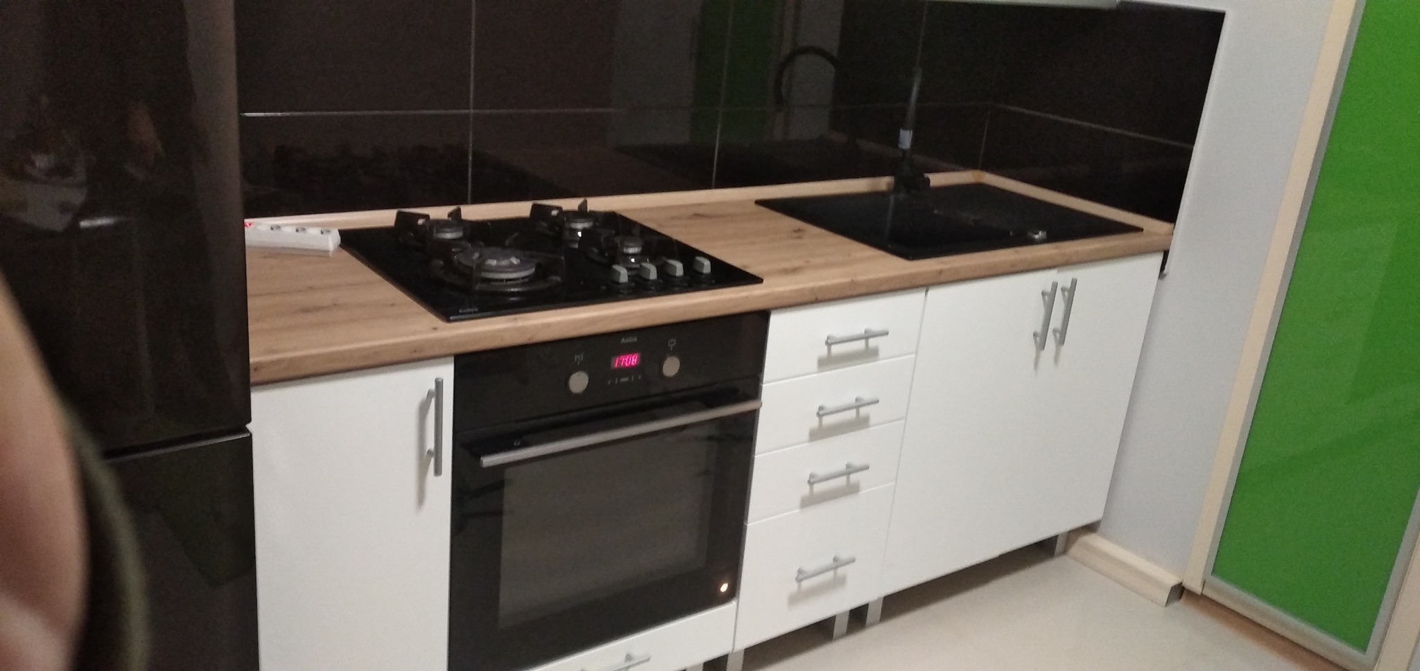 Fragment zabudowy kuchennej z białymi szafkami, blatem imitującym drewno, czarną płytą grzewczą i piekarnikiem; w tle czarne płytki na ścianie i zielone drzwi.