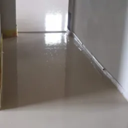 Świeżo wylana, gładka posadzka w jasnym odcieniu, odbijająca światło, z widoczną listwą przypodłogową wzdłuż ściany i przejściem do kolejnego pomieszczenia w tle.