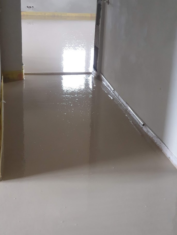 Świeżo wylana, gładka posadzka w jasnym odcieniu, odbijająca światło, z widoczną listwą przypodłogową wzdłuż ściany i przejściem do kolejnego pomieszczenia w tle.