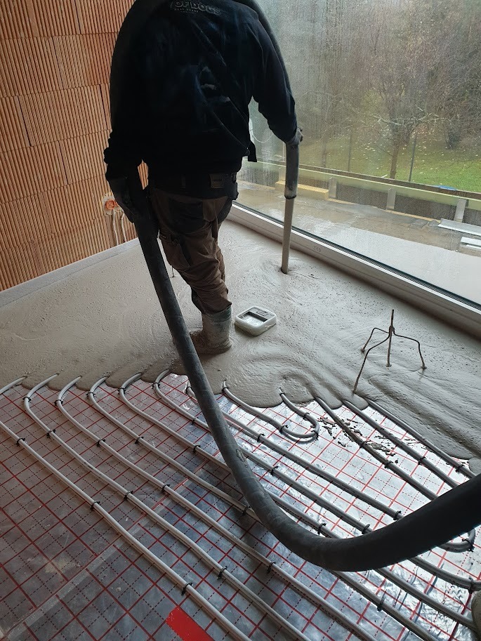 Aplikacja płynnej posadzki na ogrzewanie podłogowe w surowym stanie, widoczny pracownik z wężem i narzędzia, surowe ściany z cegły.