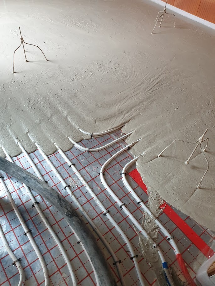 Świeżo wylana, szara masa na podłodze z widocznym systemem ogrzewania podłogowego, częściowo zakrytym, z metalowymi stojakami stabilizującymi.