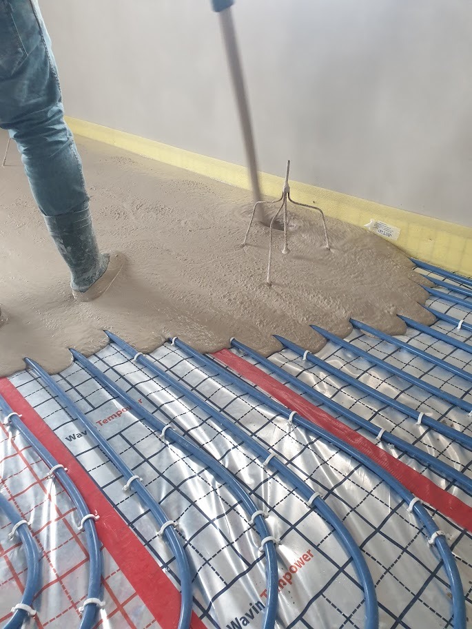 Wylewka samopoziomująca rozprowadzana nad systemem ogrzewania podłogowego, widoczny pracownik w gumowych butach i narzędzie do rozprowadzania masy.