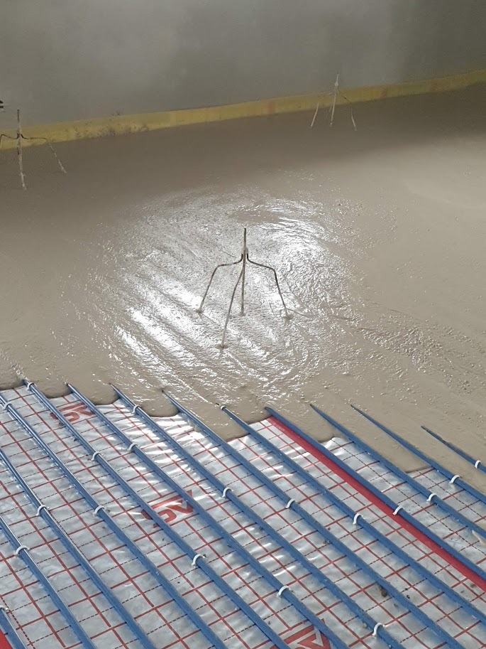 Świeżo wylana, płynna masa na systemie ogrzewania podłogowego, z widocznymi metalowymi statywami do poziomowania w tle.