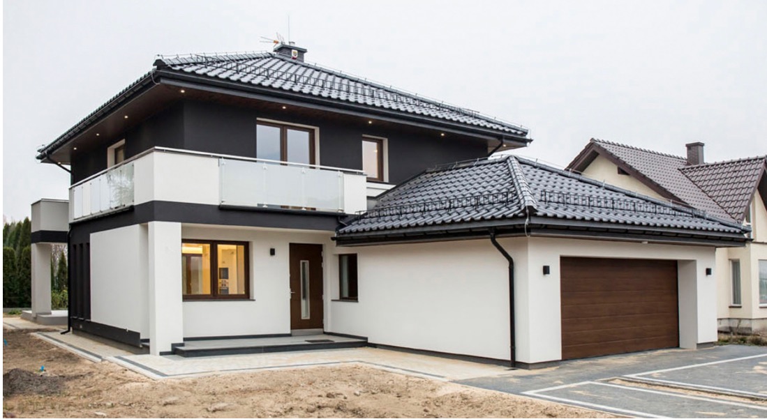 Dwukondygnacyjny dom murowany z balkonem ze szklaną balustradą, garażem z brązową bramą segmentową i czarnym dachem z dachówki ceramicznej, w trakcie wykańczania terenu wokół budynku.