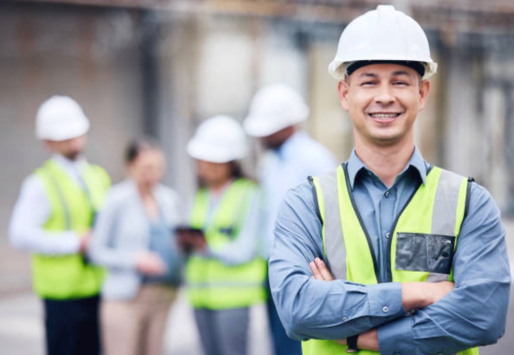 Uśmiechnięty inżynier budowlany w kasku i kamizelce odblaskowej na tle zespołu omawiającego projekt budowlany.