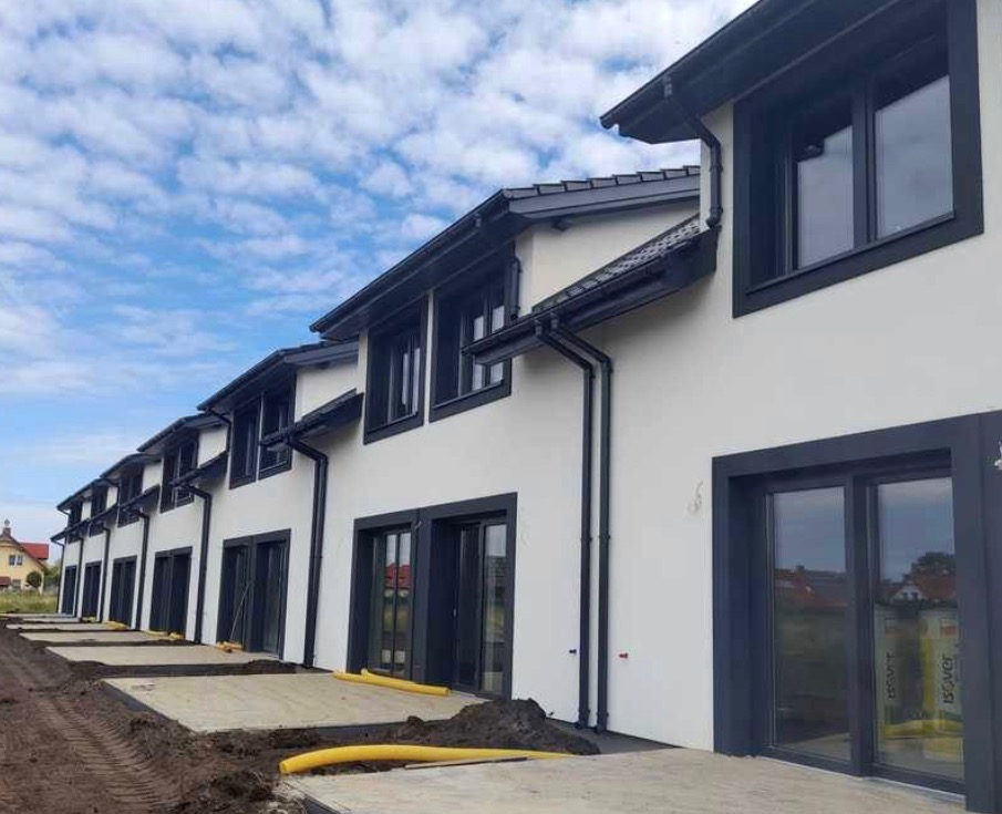 Szereg identycznych domów w stanie deweloperskim z białymi elewacjami i antracytowymi ramami okiennymi oraz rynnami, widok z poziomu gruntu na tle błękitnego nieba z chmurami.