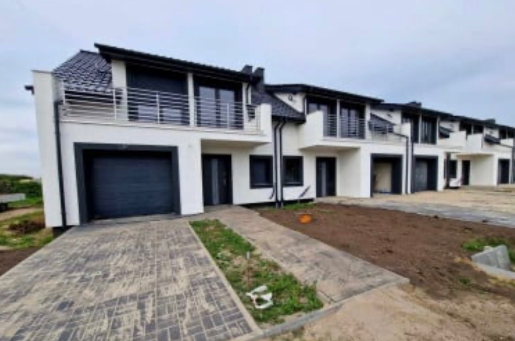 Szeregówka nowoczesnych domów z białymi elewacjami, czarnymi dachami i grafitowymi bramami garażowymi, widok z zewnątrz na podjazd wyłożony kostką.