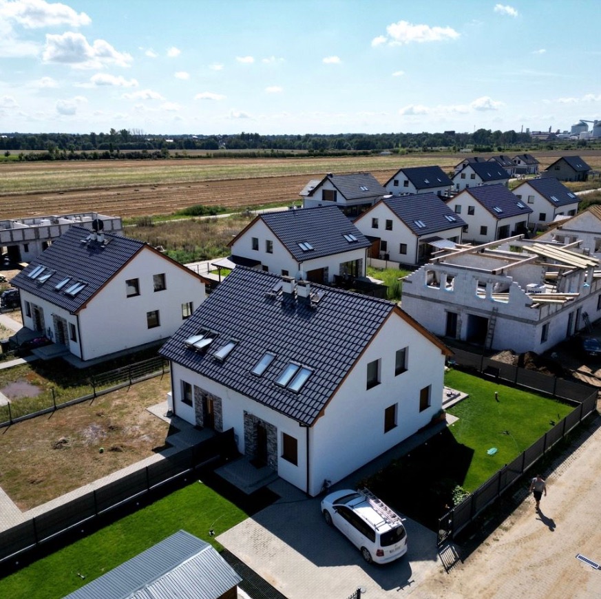 Nowoczesne osiedle domów jednorodzinnych z białymi elewacjami i grafitową dachówką, widok z lotu ptaka, jeden dom w stanie surowym, samochód dostawczy z drabiną na dachu zaparkowany na podjeździe.
