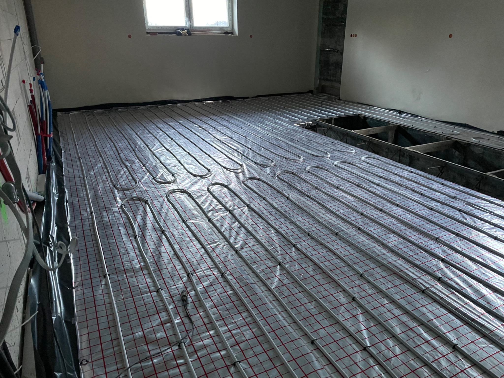 Instalacja ogrzewania podłogowego: rury grzewcze ułożone na folii z siatką w surowym wnętrzu, widoczne elementy instalacji wodnej na ścianie.