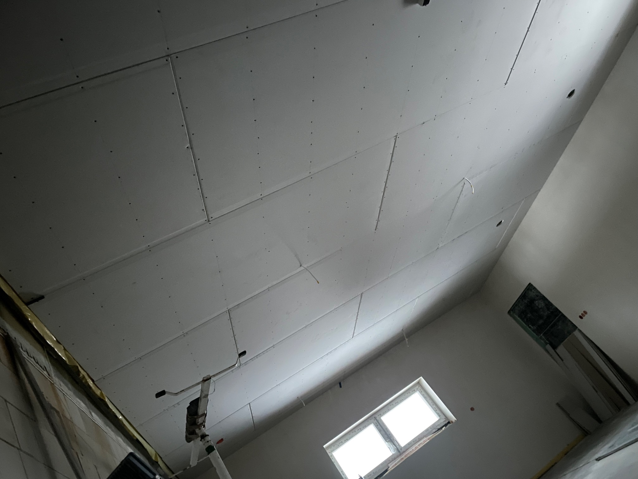 Sufit z płyt gipsowo-kartonowych w trakcie montażu, widoczne łączenia płyt i ślady po wkrętach, drabina w rogu pomieszczenia, okno w ścianie bocznej.