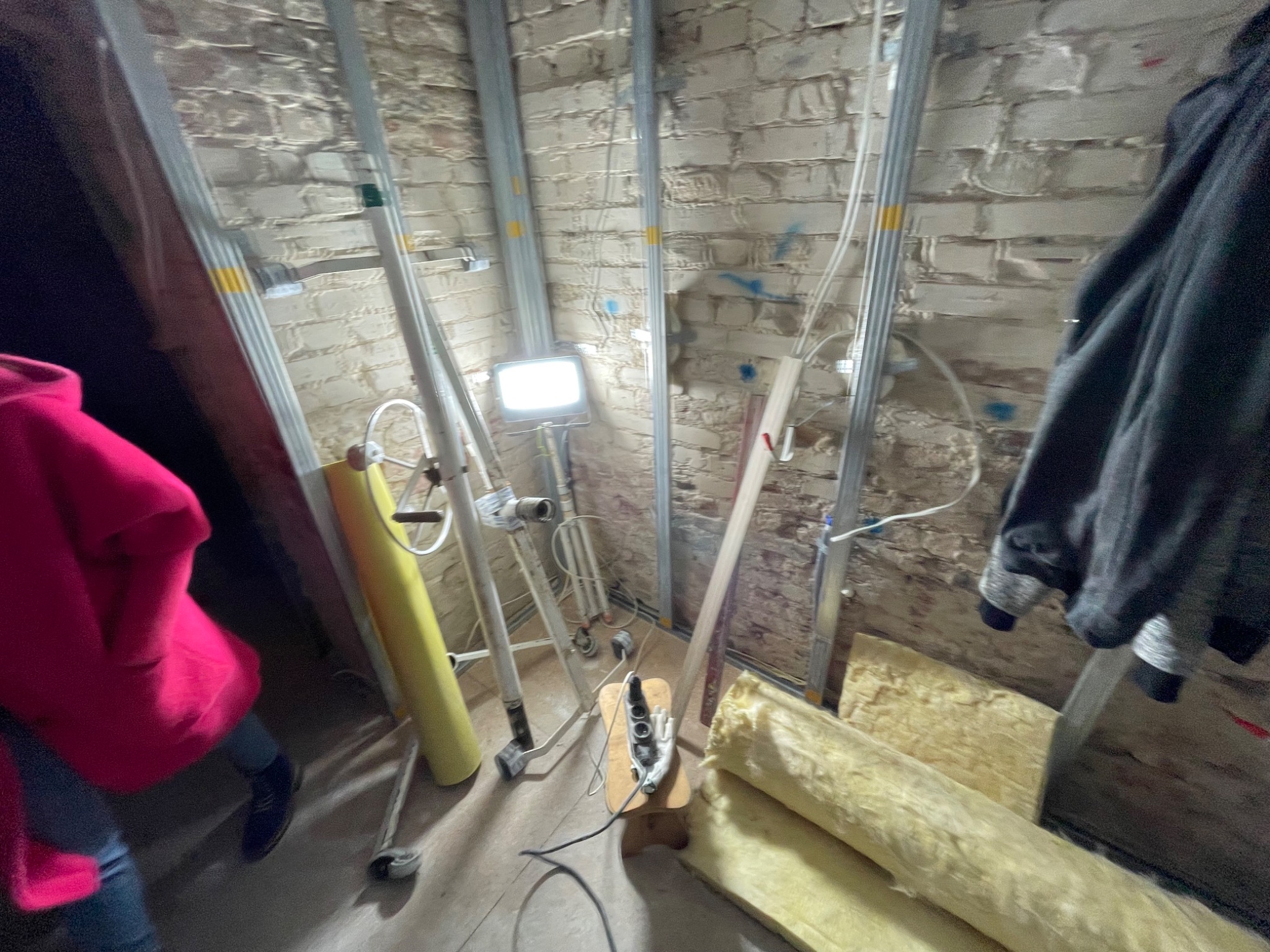 Poddasze w trakcie ocieplania: widoczne metalowe profile, fragment ściany z cegły, izolacja z wełny mineralnej, lampa budowlana i osoba w różowej bluzie.