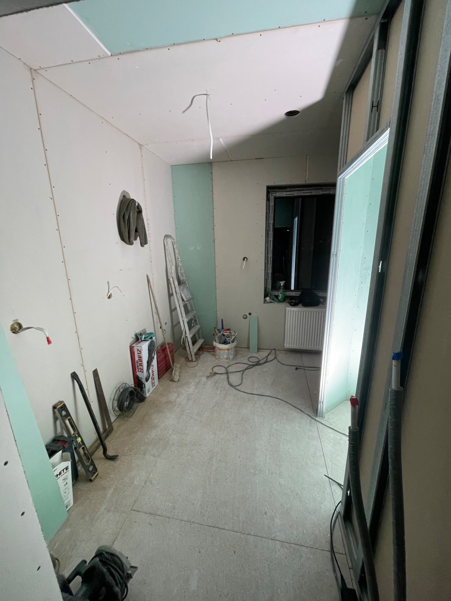 Pokój w trakcie prac wykończeniowych, widoczne płyty gipsowo-kartonowe na ścianach i suficie, metalowe profile, drabina, narzędzia i materiały budowlane na podłodze.