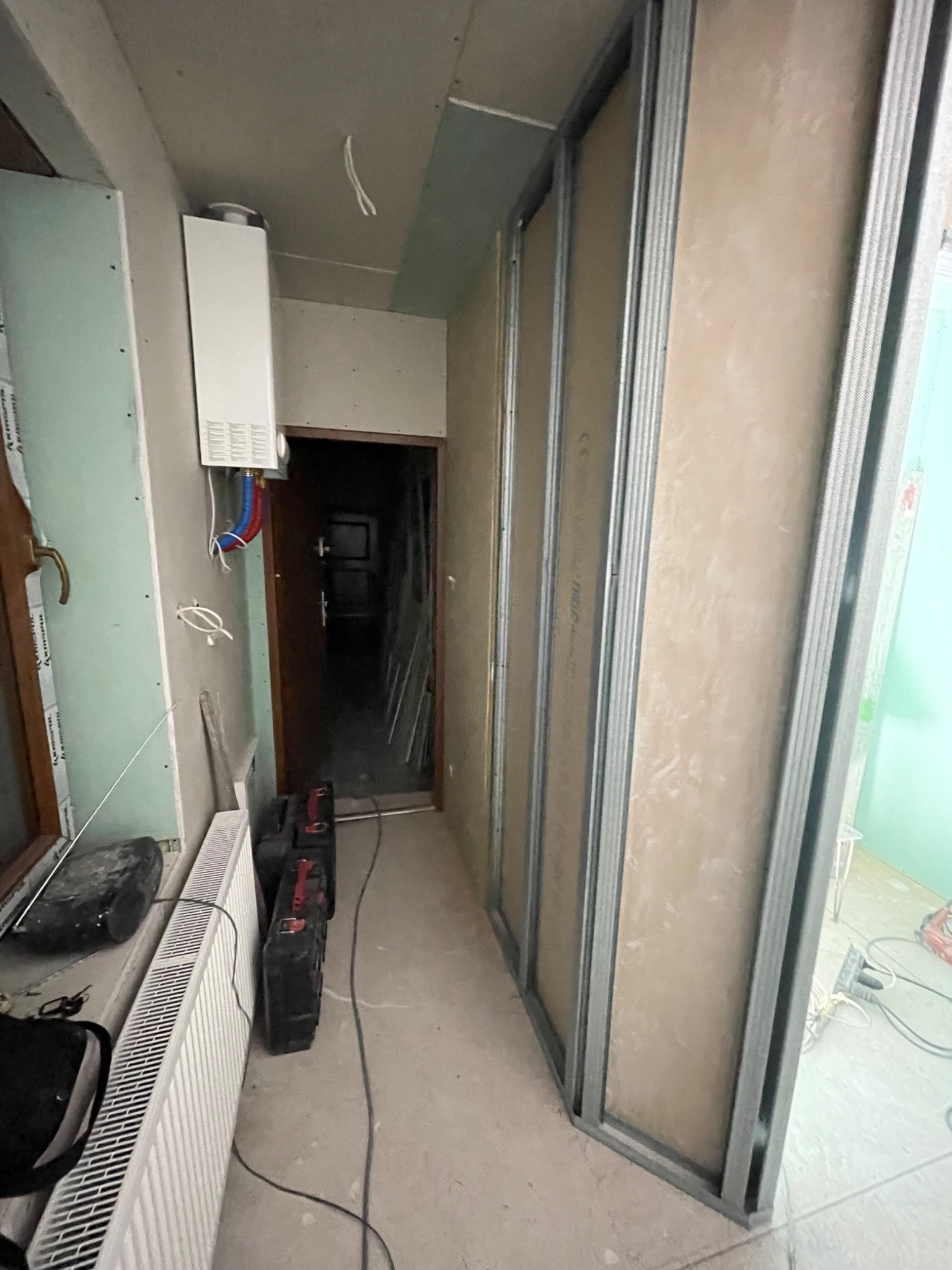 Wnętrze w trakcie remontu: widoczna metalowa konstrukcja ścian działowych z płytami kartonowo-gipsowymi, grzejnik, wiszący piec gazowy i narzędzia w tle otwartego przejścia do innego pomieszczenia.