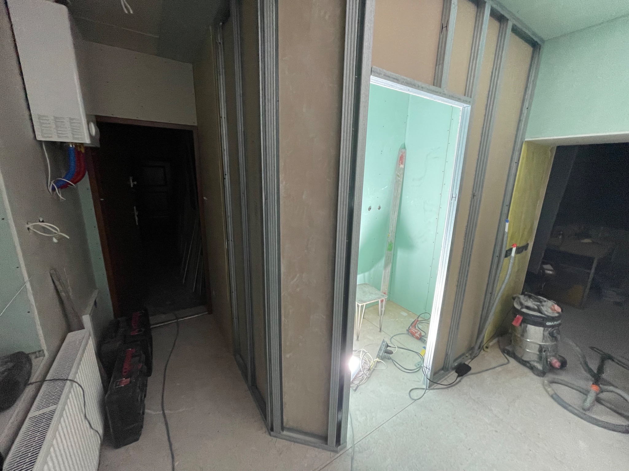 Wnętrze w trakcie prac: metalowy szkielet ścian działowych z zamontowanymi płytami gipsowo-kartonowymi, widoczny grzejnik, narzędzia i odkurzacz przemysłowy.