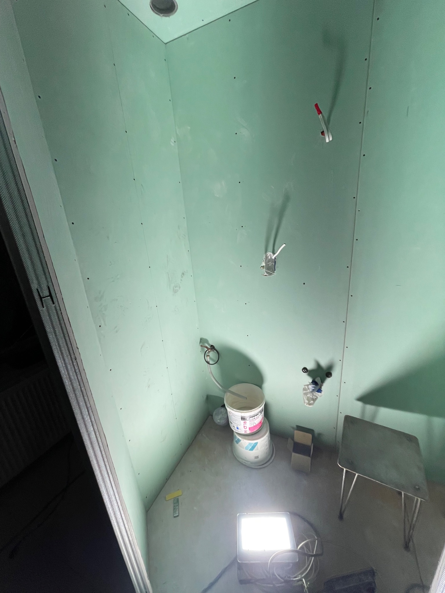 Wnętrze łazienki w trakcie prac: ściany wyłożone zielonymi płytami gipsowo-kartonowymi, widoczne puszki elektryczne, wiaderka z masą szpachlową, przenośna lampa halogenowa oświetlająca podłogę.