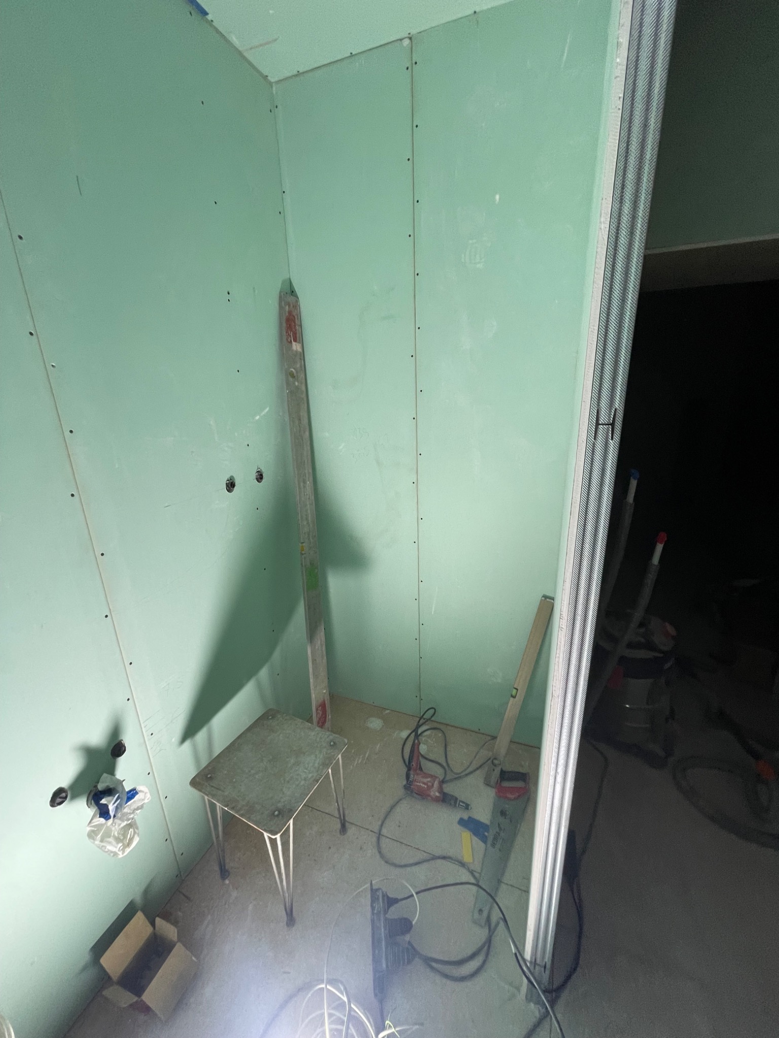 Wnętrze w trakcie prac wykończeniowych: zielone płyty gipsowo-kartonowe na ścianach, widoczne otwory na gniazdka elektryczne, poziomica oparta o ścianę, stołek, narzędzia budowlane i metalowy...