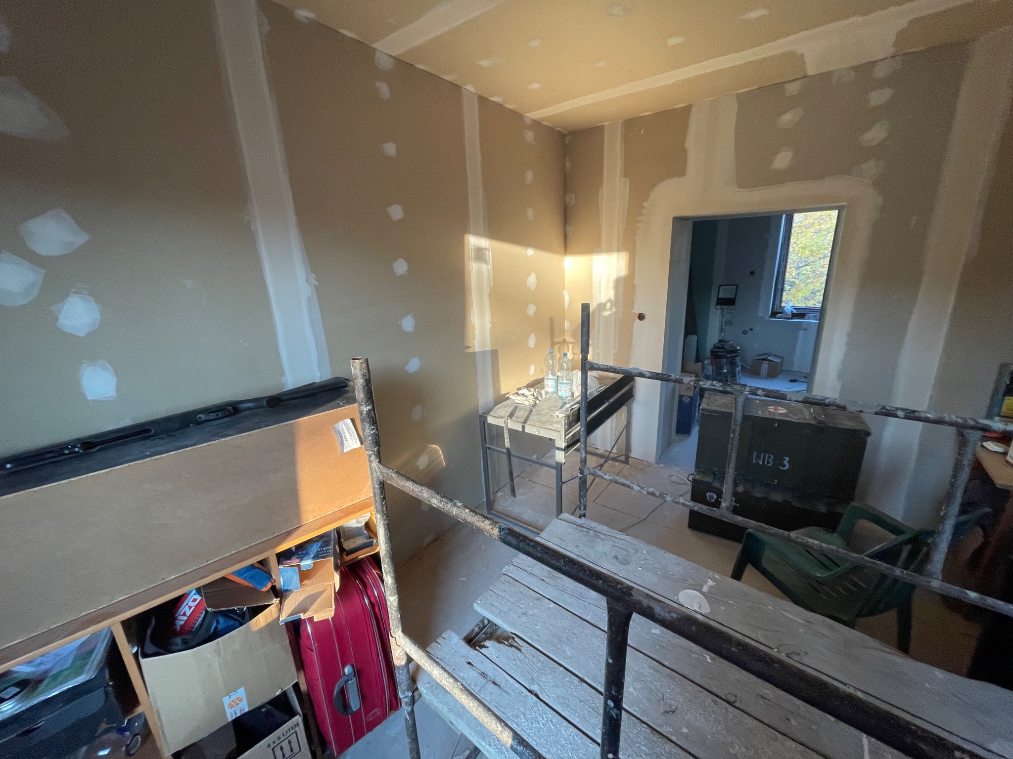 Pokój w trakcie prac wykończeniowych, widoczne ściany z płyt gipsowo-kartonowych ze śladami szpachlowania, rusztowanie w pomieszczeniu, kartony i walizka w tle, otwarte przejście do kolejnego...