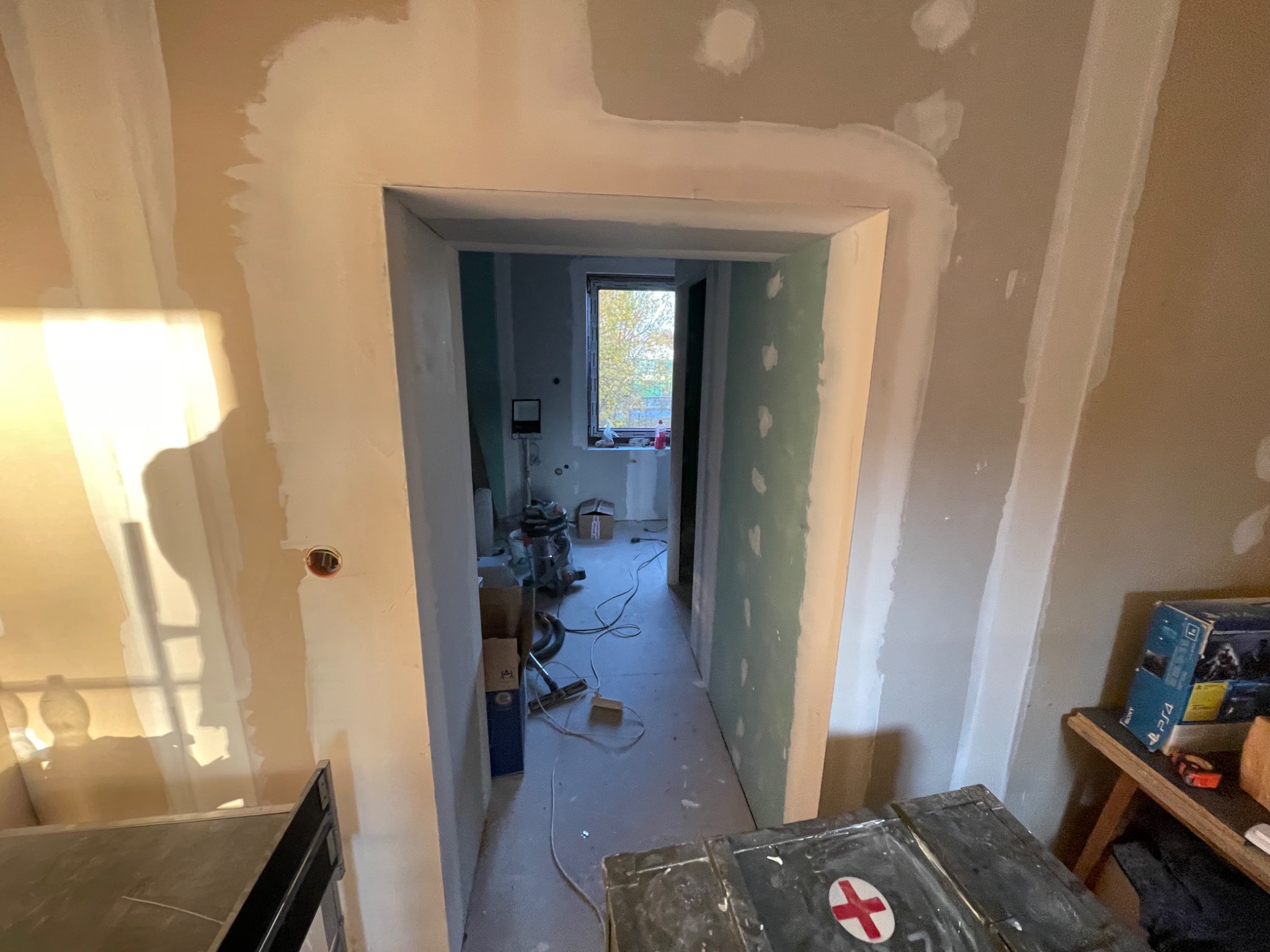 Wnętrze w trakcie prac przygotowawczych: ściany po szpachlowaniu, widoczna brama drzwiowa z nałożoną warstwą gładzi, narzędzia i materiały budowlane w tle.