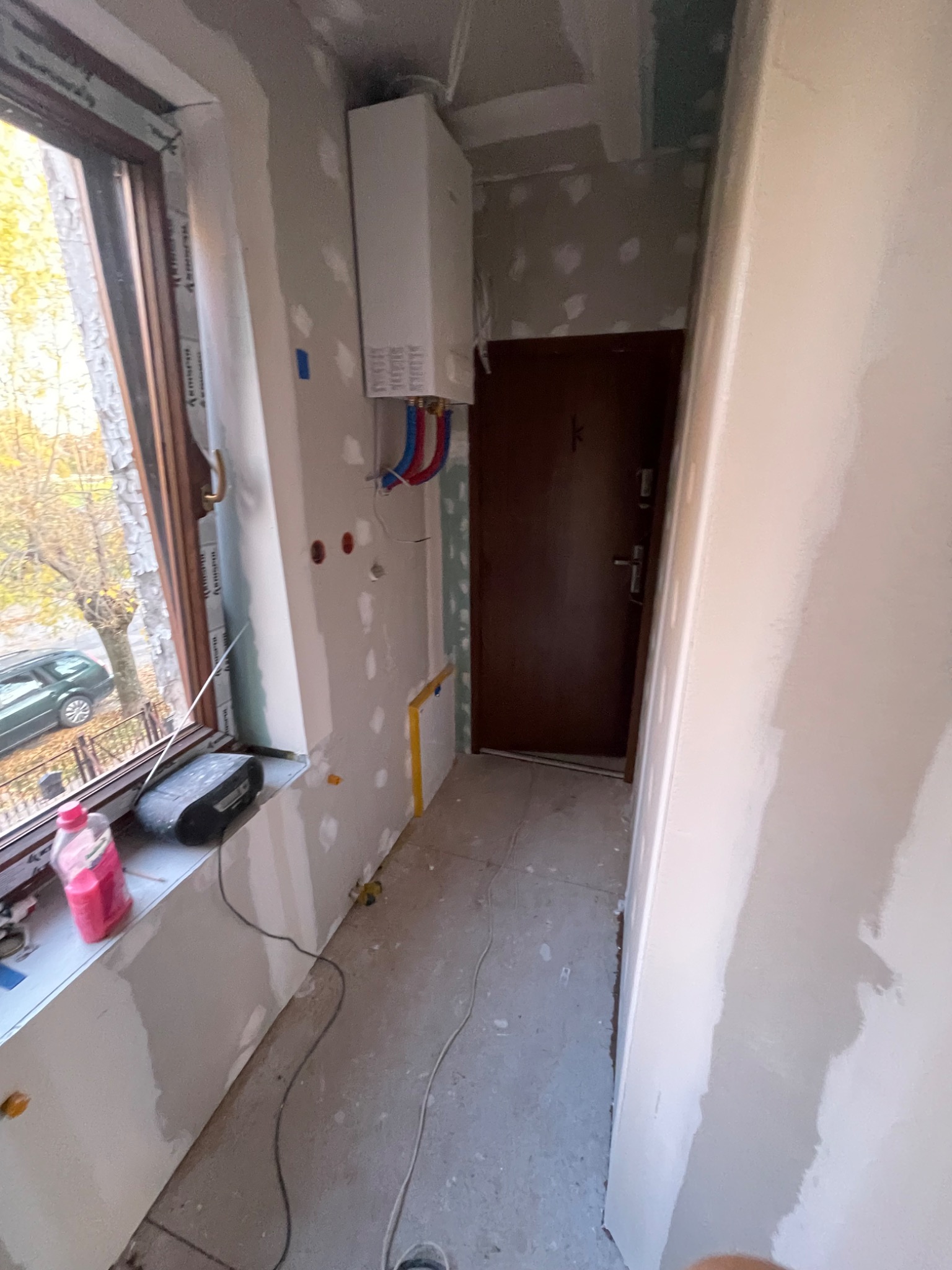 Korytarz w trakcie prac wykończeniowych; ściany przygotowane do malowania, widoczne ślady szpachlowania, grzejnik na ścianie z podłączonymi rurami, okno z widokiem na drzewo i samochód.