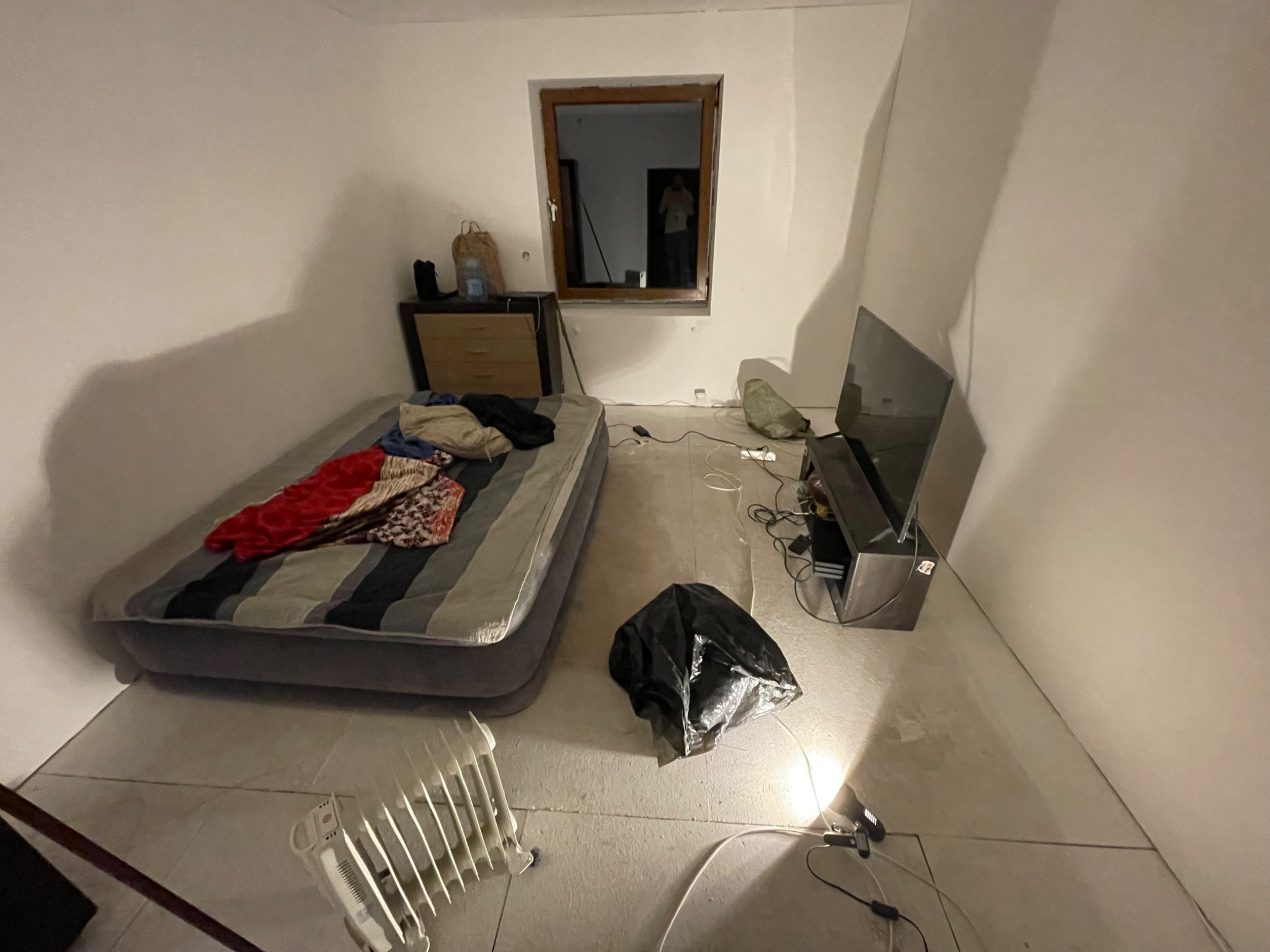 Pokój w trakcie remontu: materac dmuchany z pościelą, grzejnik elektryczny, telewizor na szafce RTV, okno z widocznym odbiciem osoby, oraz czarna torba na gruz na podłodze.