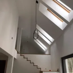 Wnętrze w trakcie wykańczania: gładkie, białe ściany, drewniane schody bez balustrady, okna dachowe w skośnym suficie i wisząca lampa na kablu.