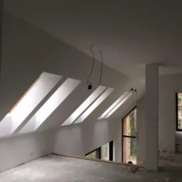 Wnętrze poddasza w trakcie wykańczania: białe ściany, okna dachowe w skosie, surowa podłoga, widoczne kable i elementy instalacji elektrycznej, duże okno z widokiem na las.