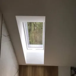 Poddasze z oknem dachowym, widok na drzewa, ściany wykończone płytkami imitującymi marmur i panelami drewnopodobnymi, fragment surowej ściany.