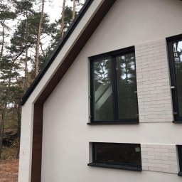 Krawczyk-bud - Nowoczesny budynek z elewacją w odcieniach bieli i szarości, z elementami dekoracyjnymi imitującymi cegłę i czarnymi ramami okiennymi, na tle drzew.