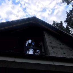 Krawczyk-bud - Niski kąt widzenia na szczyt domu w trakcie prac elewacyjnych, widoczne płyty izolacyjne z pozostałościami zaprawy, okno z refleksami drzew i nieba, oraz fragment dachu z ciemną dachówką.