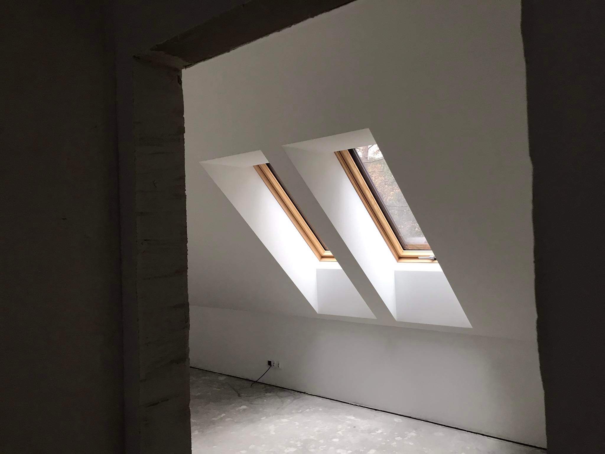 Poddasze po remoncie z dwoma oknami dachowymi, ściany świeżo pomalowane na biało, widoczne gniazdka elektryczne przy podłodze, surowa ościeżnica drzwiowa w tle.