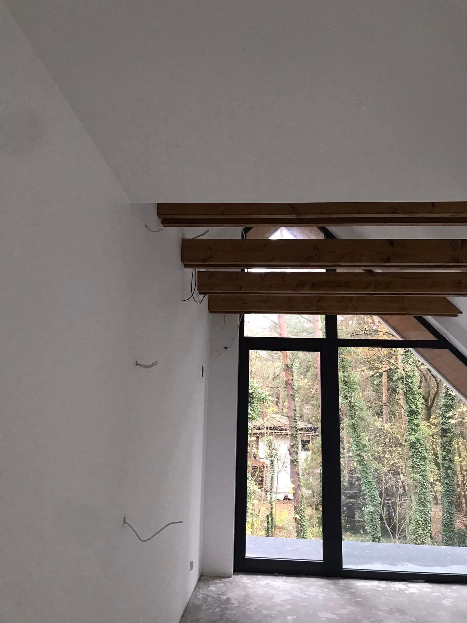 Pokój na poddaszu w trakcie wykańczania: ściany po nałożeniu gładzi, widoczne drewniane elementy konstrukcyjne stropu i okno z widokiem na las.