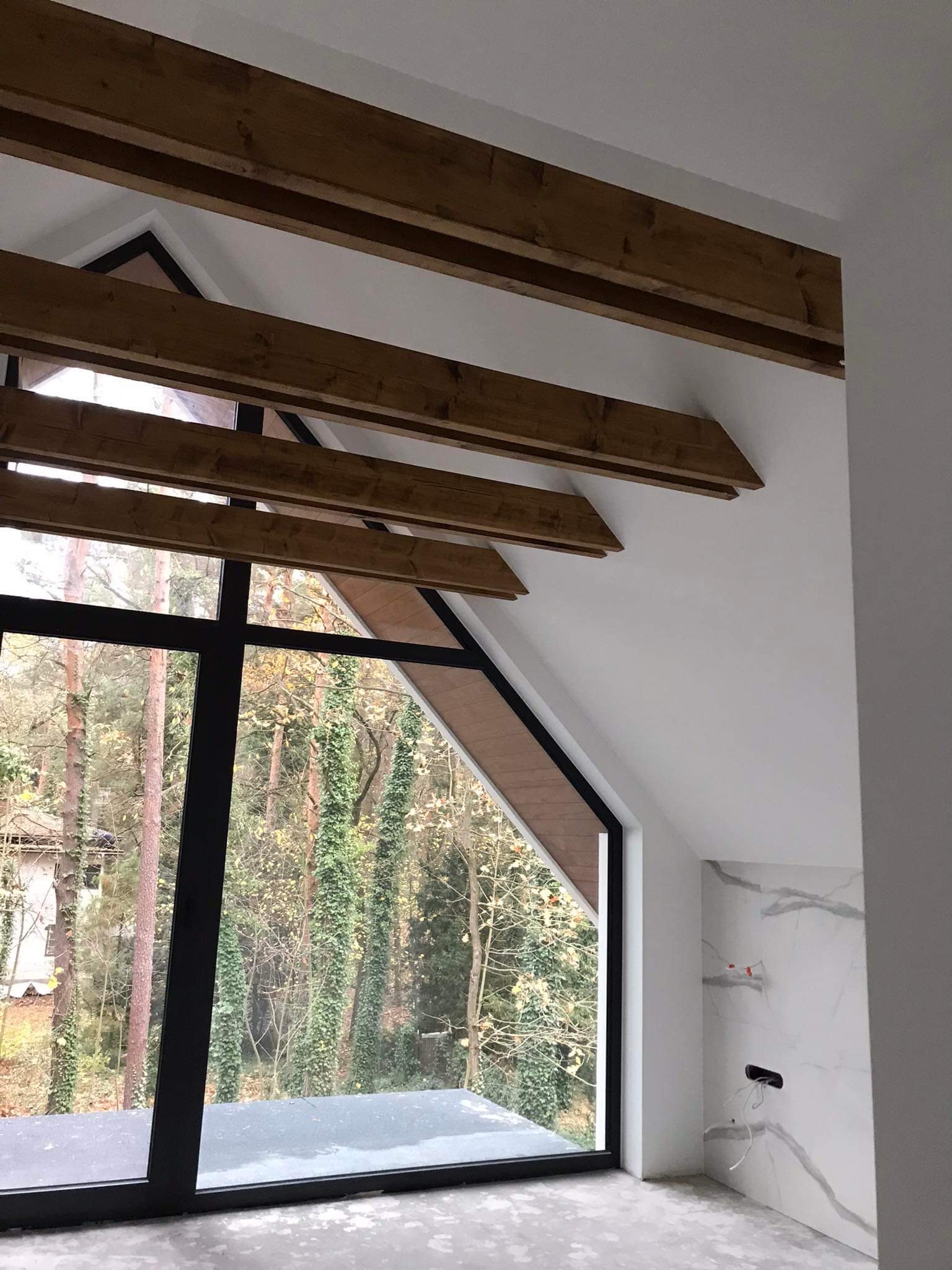 Wnętrze poddasza z drewnianymi belkami stropowymi, dużym oknem z widokiem na las i ścianą wykończoną płytami imitującymi marmur z wystającymi przewodami elektrycznymi.