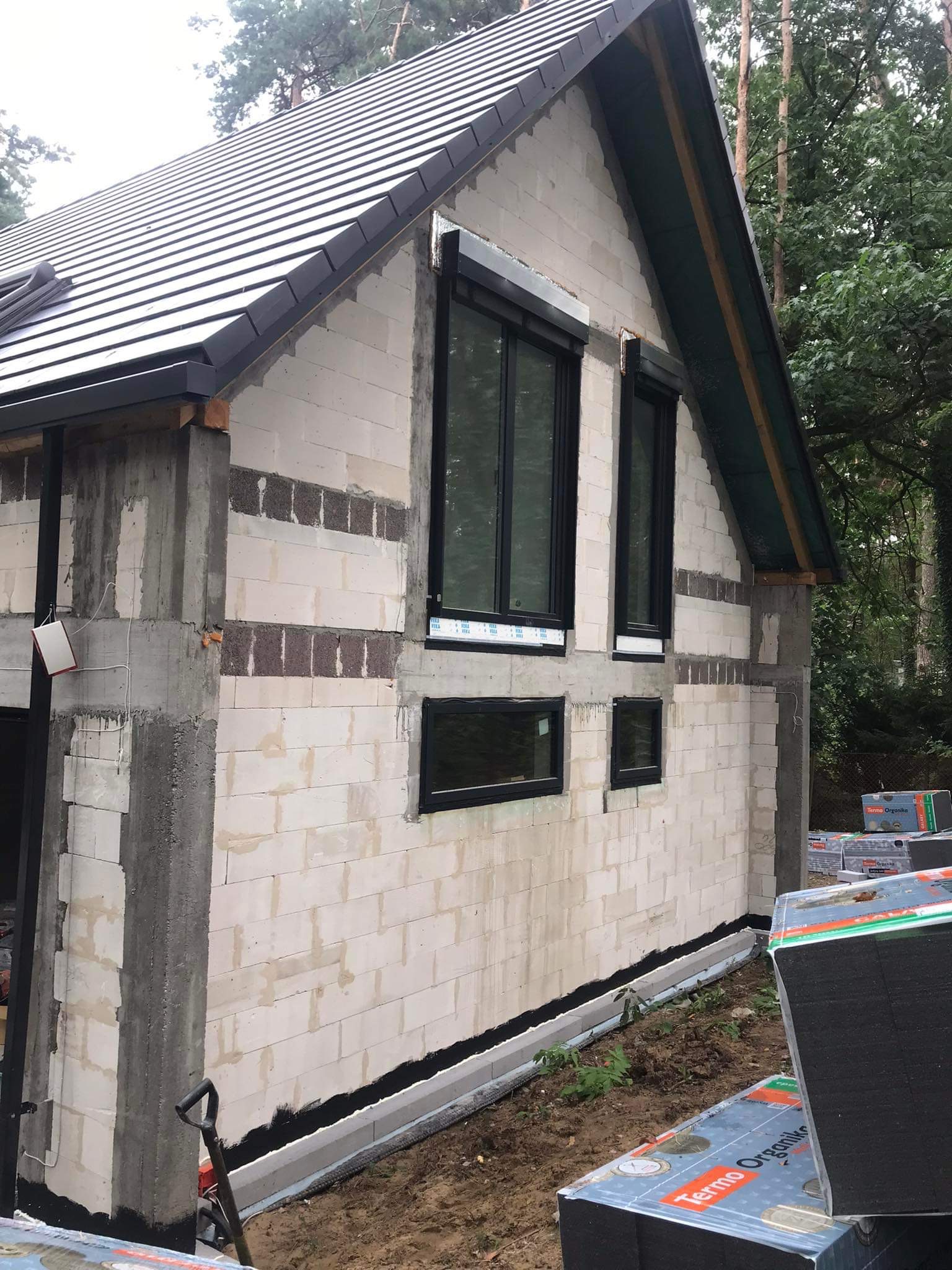 Surowy stan elewacji domu jednorodzinnego z białych bloczków z widocznymi czarnymi oknami i przygotowanymi paczkami styropianu Termo Organika, szarym fundamentem i czarną izolacją przeciwwilgociową.