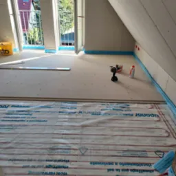 Poddasze w trakcie prac wykończeniowych: widoczne płyty Fermacell na podłodze, system ogrzewania podłogowego Uponor, narzędzia (wiertarka), otwarte okna balkonowe w skosach dachu.