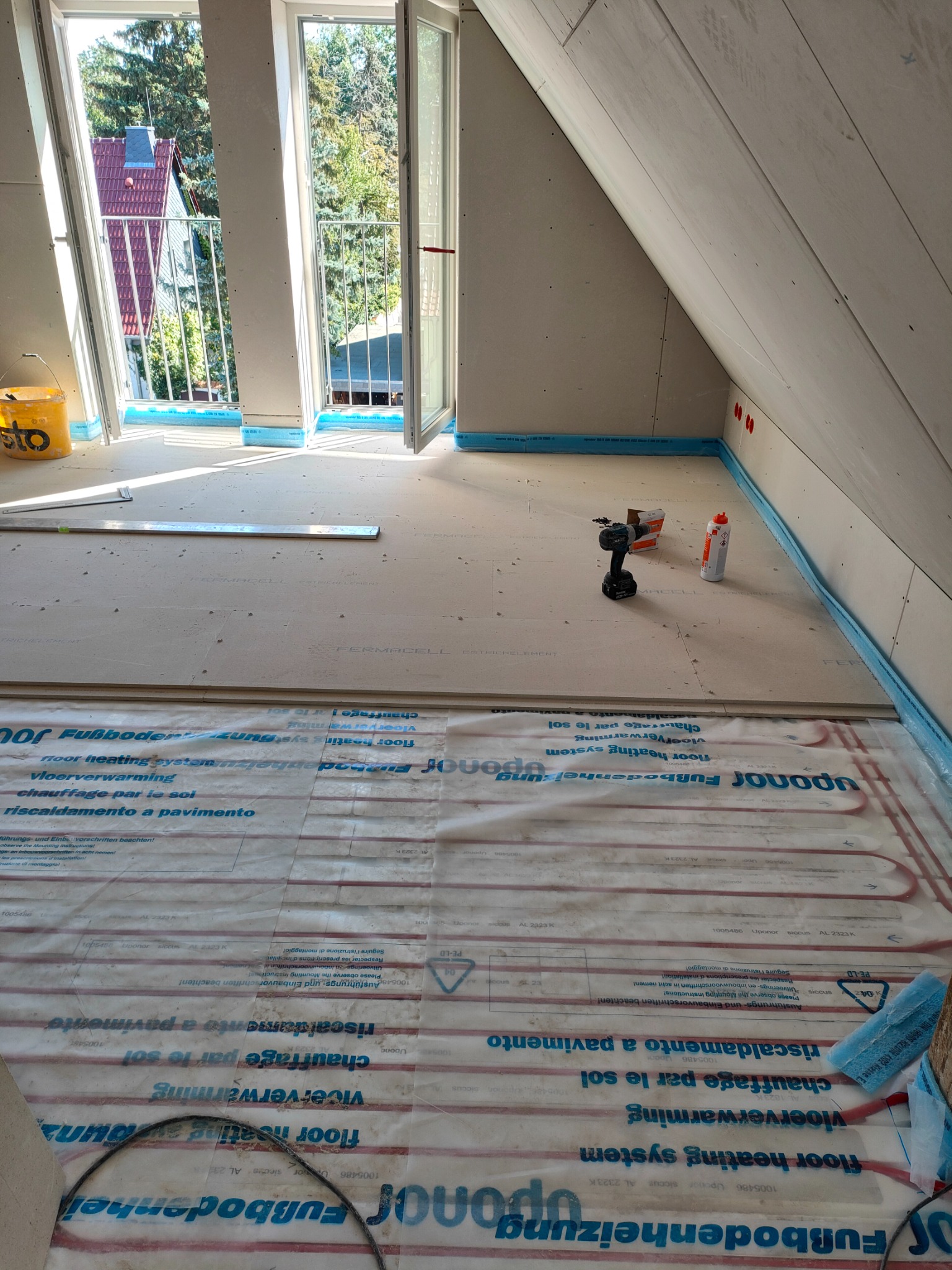 Poddasze w trakcie prac wykończeniowych: widoczne płyty Fermacell na podłodze, system ogrzewania podłogowego Uponor, narzędzia (wiertarka), otwarte okna balkonowe w skosach dachu.