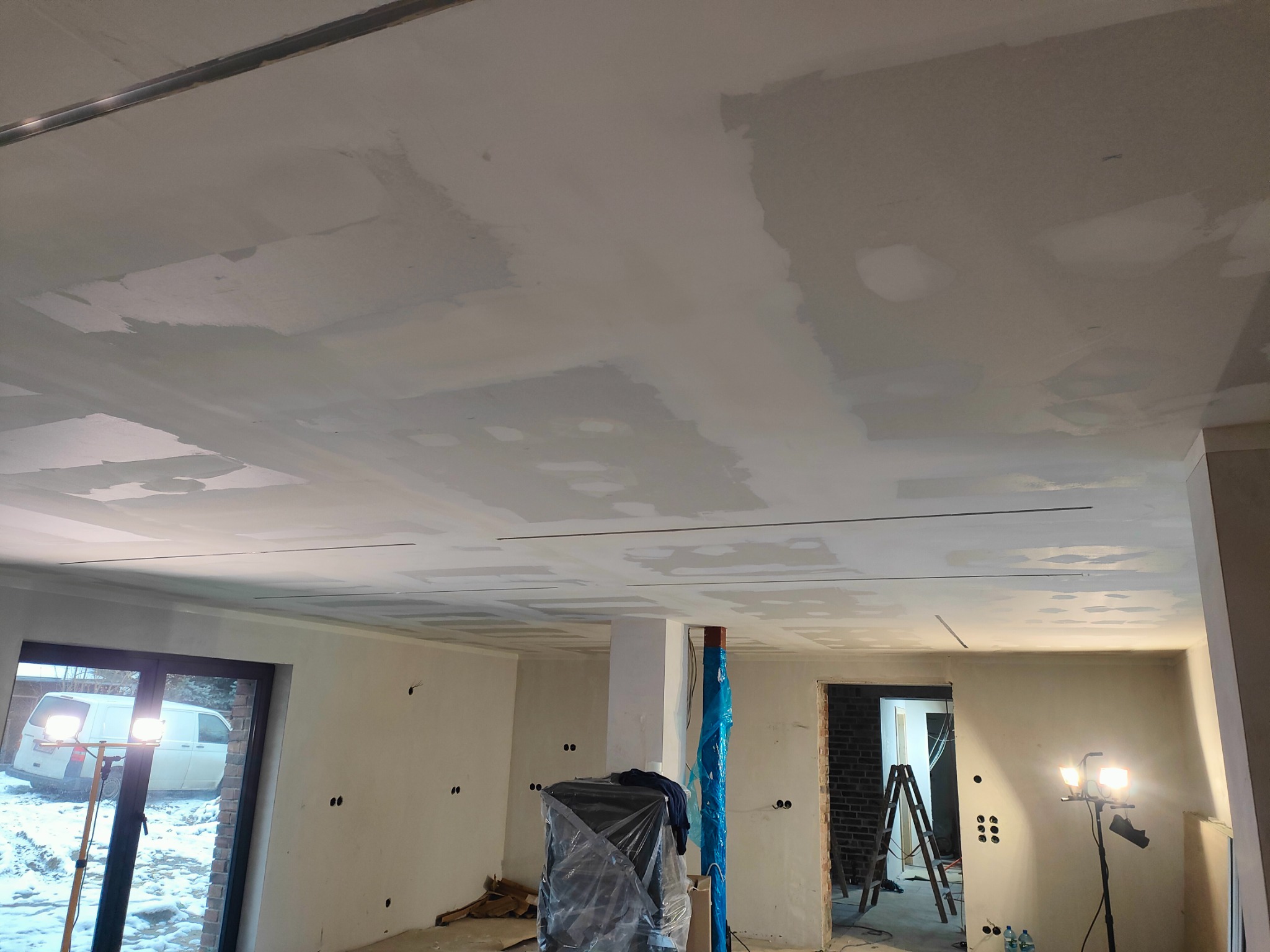 Wnętrze w trakcie remontu: sufit z płyt kartonowo-gipsowych z widocznymi łączeniami przed szpachlowaniem, reflektory budowlane oświetlają pomieszczenie, okno z widokiem na zimowy krajobraz...