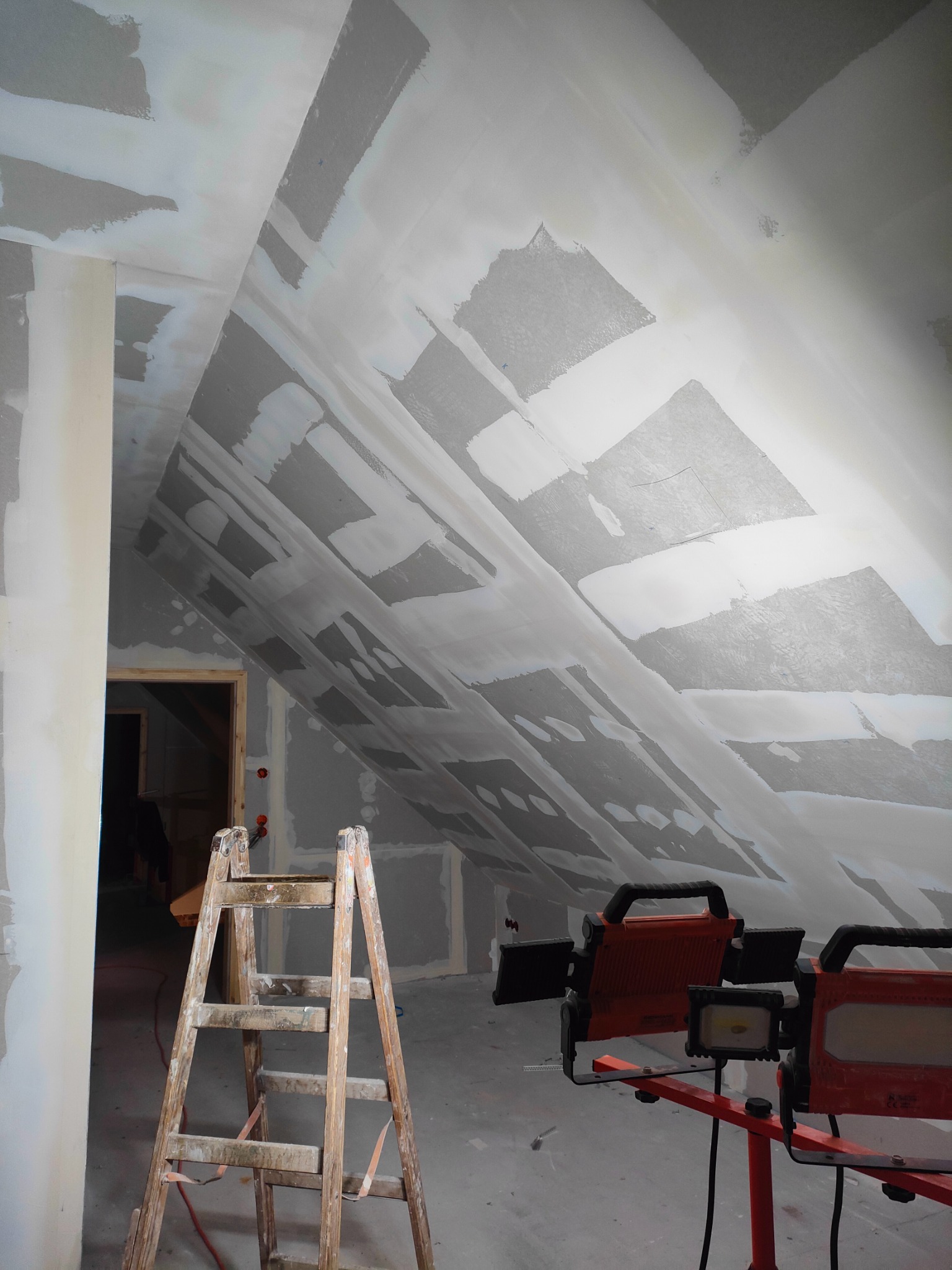 Poddasze w trakcie prac wykończeniowych, widoczne płyty kartonowo-gipsowe na skośnych ścianach, częściowo pokryte warstwą szpachli, drewniana drabina oparta o ścianę oraz lampy budowlane...