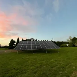 Panel słoneczny na trawniku przed domem, widok na zachodzące słońce barwiące niebo na różowo.