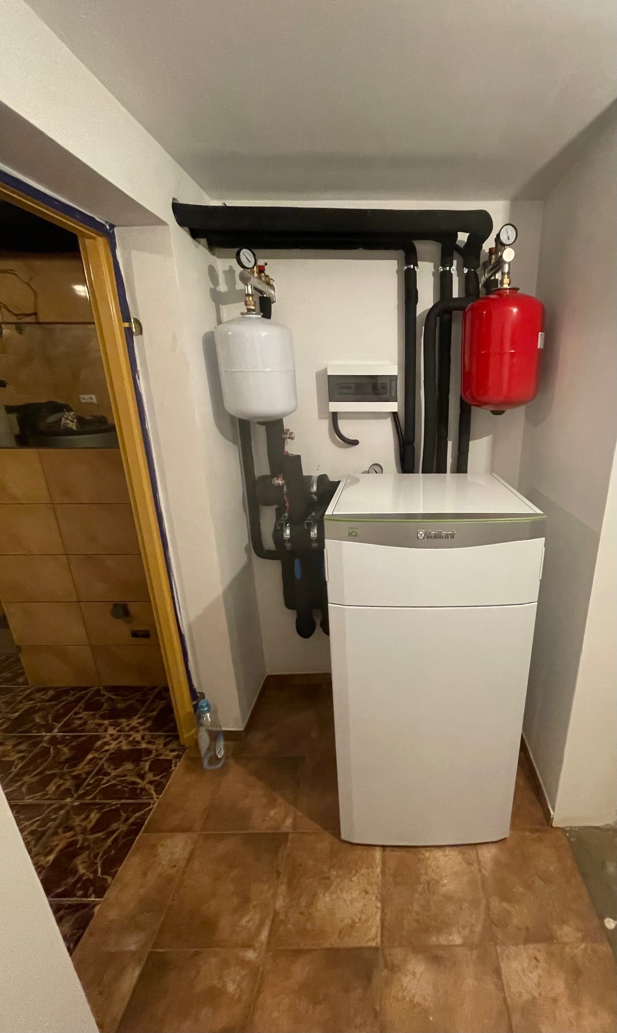 Biała jednostka wewnętrzna z czerwonym i białym zbiornikiem wyrównawczym, czarne rury instalacyjne, brązowe płytki podłogowe, plastikowa butelka po wodzie w rogu pomieszczenia.