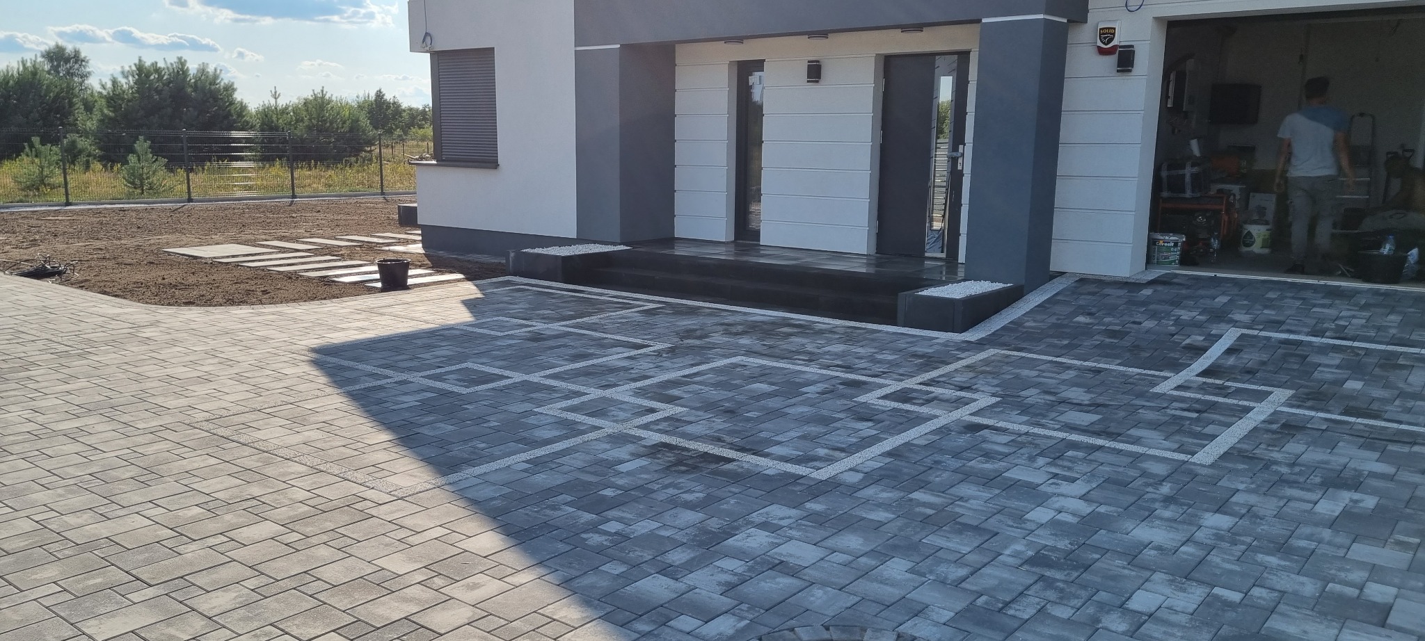 Ułożona kostka brukowa w geometryczny wzór przed nowoczesnym domem z szarą elewacją, widoczny fragment trawnika z płytami chodnikowymi i otwarty garaż z osobą w środku.