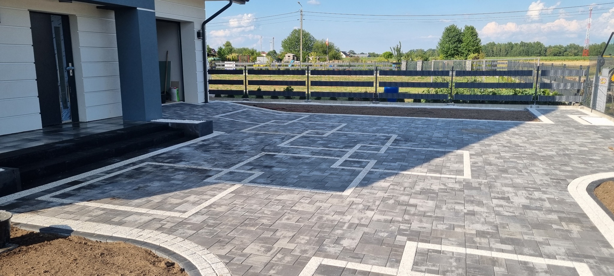 Ułożona szara kostka brukowa z białymi wstawkami tworzącymi geometryczny wzór przed nowoczesnym domem z szarą stolarką okienną, widok na posesję z czarnym płotem panelowym i zielenią w tle.