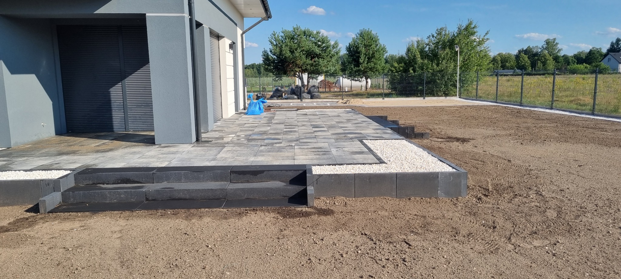 Nowoczesny taras z kostki brukowej w odcieniach szarości, z czarnymi schodami i obrzeżem z białego grysu, przylegający do budynku z szarą elewacją i roletami zewnętrznymi. W tle widoczny ogród...