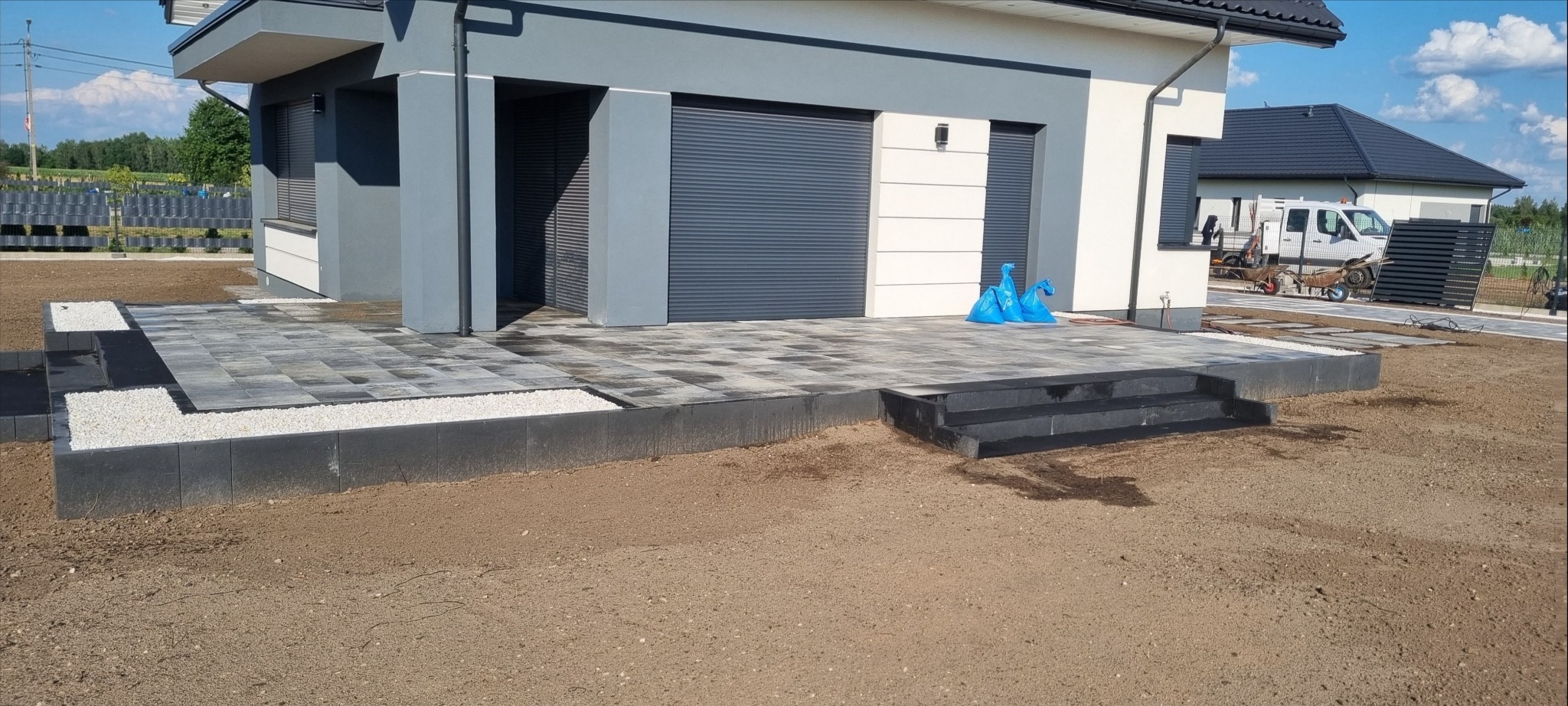 Nowoczesny taras z szarej kostki brukowej przed domem z szarą elewacją i roletami zewnętrznymi, biały żwir na obrzeżach, świeżo obsypana ziemia wokół.