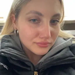 Portret młodej kobiety z blond włosami, srebrnymi kolczykami i szarą bluzą z kapturem, widoczny z bliska zamek błyskawiczny bluzy w kolorze złotym.